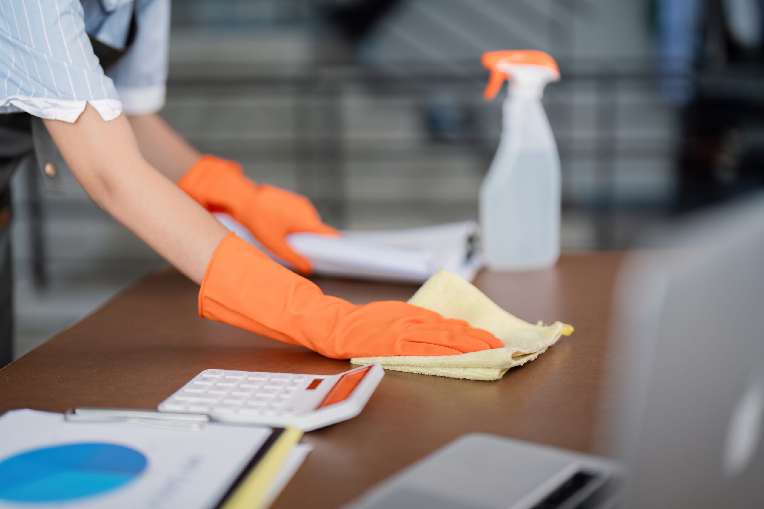 Female office cleaner wearing orange gloves, wiping a desk with cleaning supplies and documents, emphasizing workspace maintenance